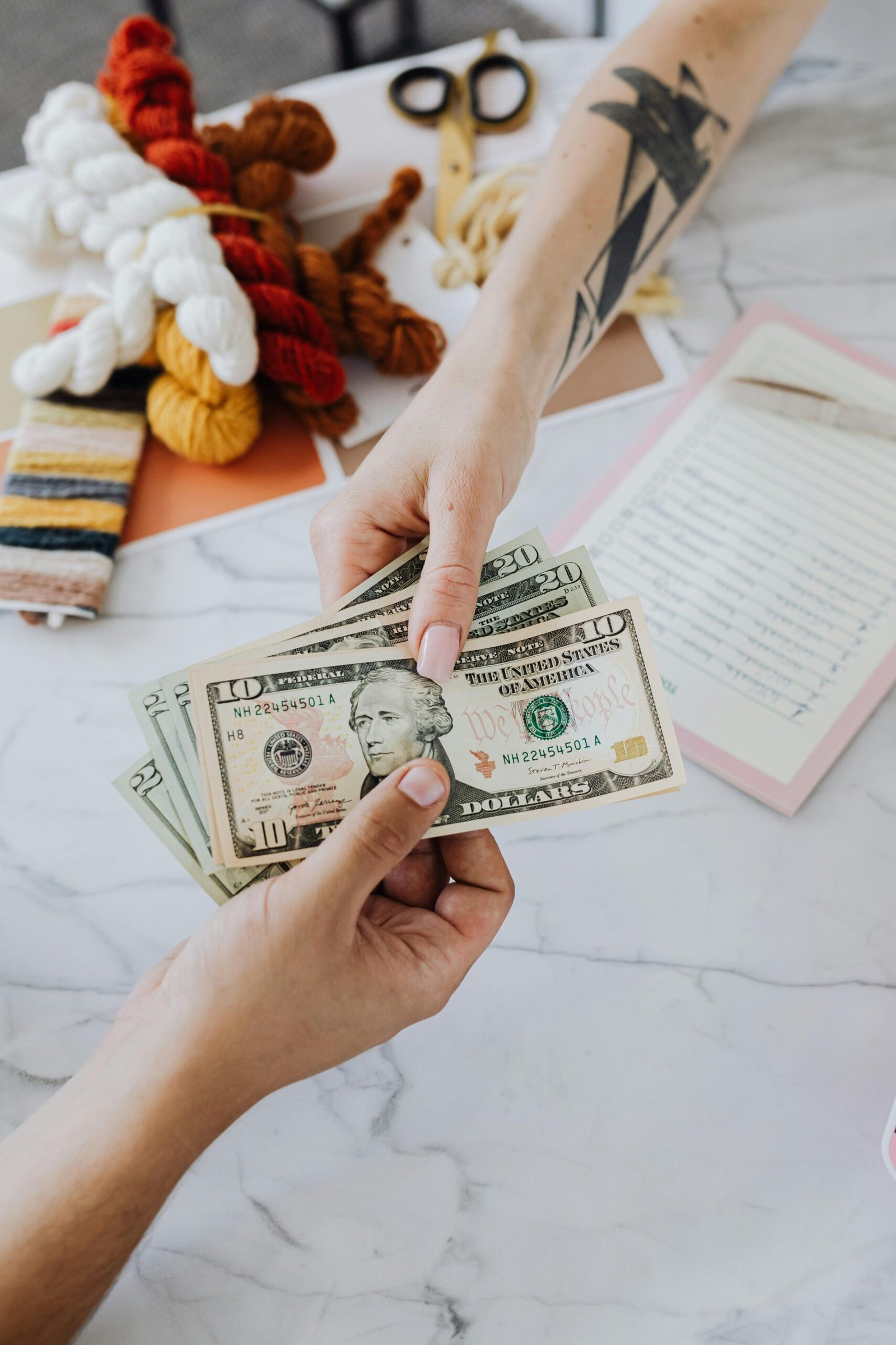 Hands exchanging US dollars on a table with crafting supplies, highlighting financial transactions.